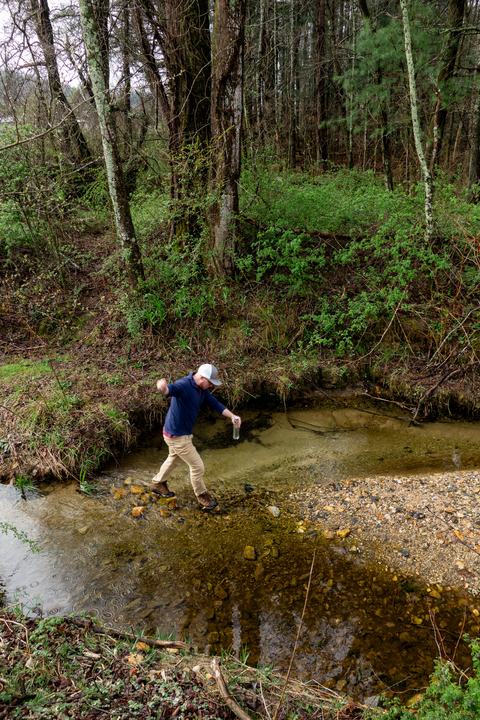 Person crossing creek with insect sample jar next to woods