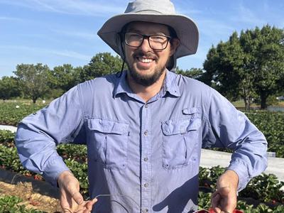 Austin Wrenn, president of the North American Strawberry Association, smiles while holding multiple red baskets of freshly picked strawberries in a farm field.