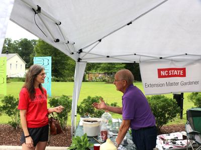 A Master Gardener volunteer staffing an information booth shares sustainable gardening advice with a community member.