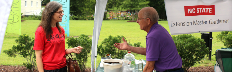 At an NC State Extension Master Gardener booth, a woman in a red shirt and an older man in a purple shirt chat under a tent in a grassy garden.