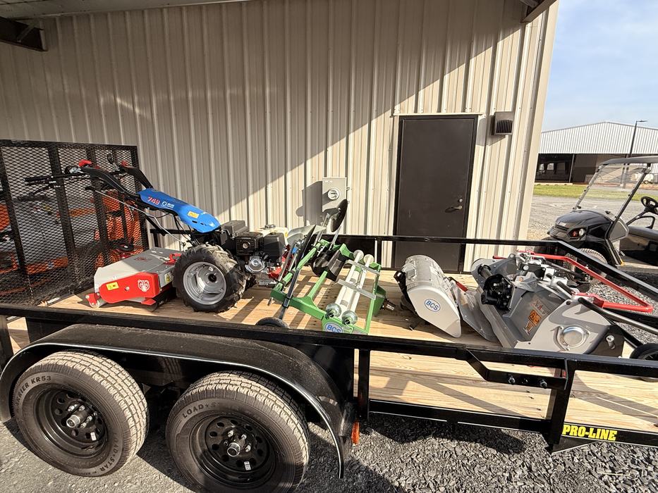 A blue BCS 749 two-wheel walk-behind tractor with various attachments, including a flail mower and a tiller, is loaded onto a black Pro-Line utility trailer.