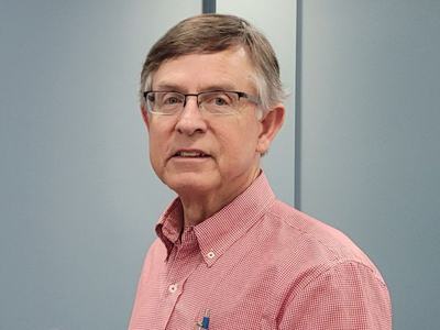 A man with light brown hair and glasses, wearing a red and white checkered button-down shirt, posing against a neutral blue-grey background