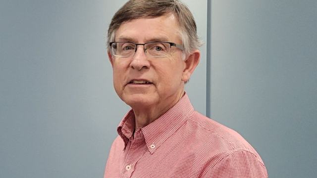 A man with light brown hair and glasses, wearing a red and white checkered button-down shirt, posing against a neutral blue-grey background