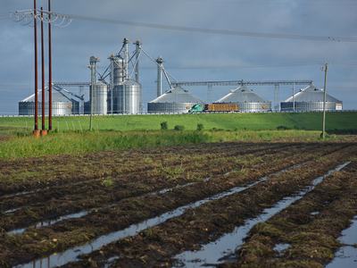 A farm landscape with wet fields, silos in the background, and stormy skies.
