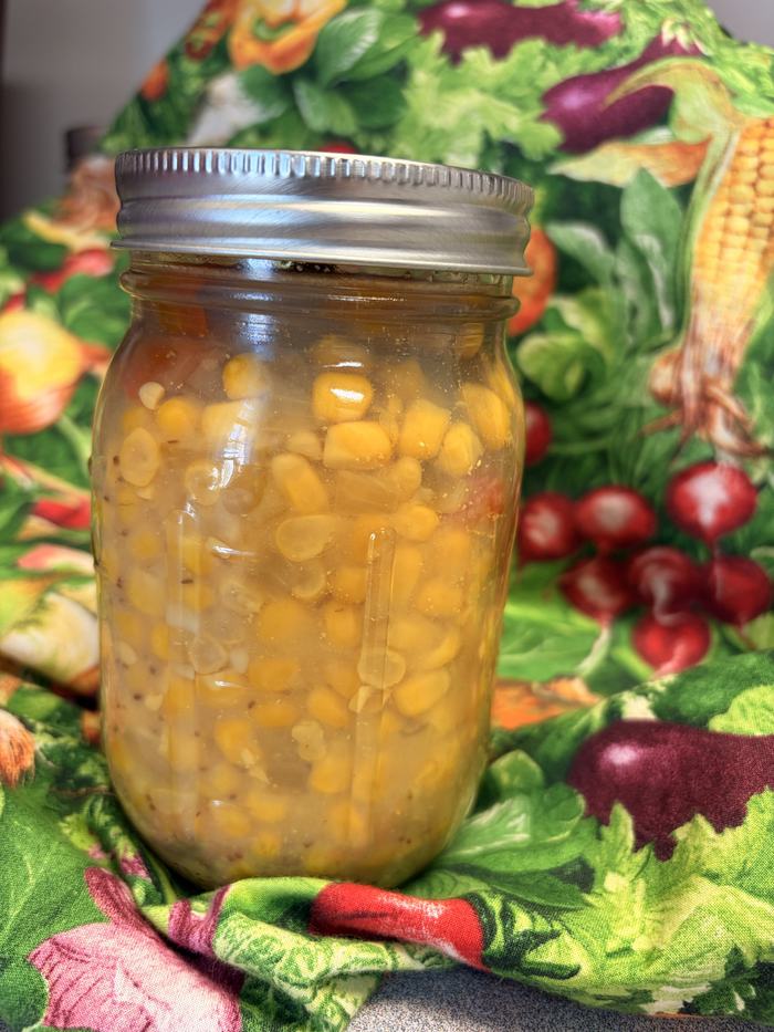 This is a vertical, close-up photograph of a single glass Mason jar filled with preserved corn. The jar is set against a colorful, busy background of fabric printed with various vegetables (radishes, corn, and eggplant).