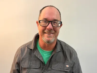 A smiling man with short brown hair, a salt-and-pepper beard and black-framed glasses wears a gray button-up shirt over a green T-shirt against a softly lit, neutral background.