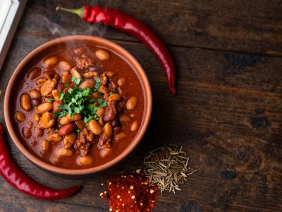 A bowl of chili flanked by two chili peppers and a pile of seasonings in front of the bowl.