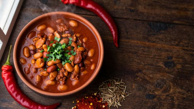 A bowl of chili flanked by two chili peppers and a pile of seasonings in front of the bowl.
