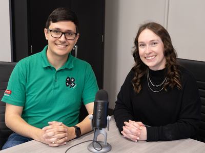 Two state 4-H staff members sit at a table with a microphone between them, smiling toward the camera before a North Carolina 4-H Clover Cast podcast recording.