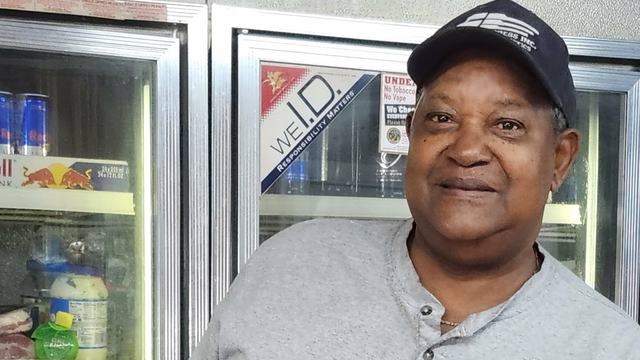 Man wearing a gray shirt and baseball cap stands in front of a refrigerated display case, with a “Small Farms Week” card displayed in front of him.