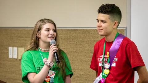 Two 4-H members during a 4-H Congress workshop: a girl in a green shirt holds a microphone and speaks, while a boy in a red shirt listens and smiles.