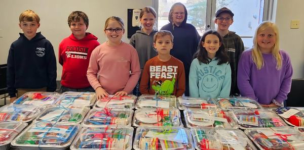 A group of seven children stands behind a long table stacked with dozens of care packages wrapped in plastic and bows. The children, smiling and varying in age, are wearing various casual outfits and hoodies.