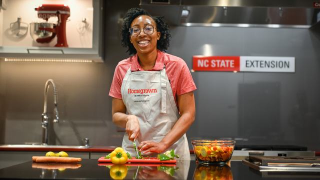 An NC State Extension agent smiles while cutting and preparing fresh vegetables, such as squash and red and green peppers, on a kitchen countertop.