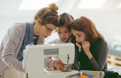 Adult woman with glasses teaching two young girls how to sew on a sewing machine.