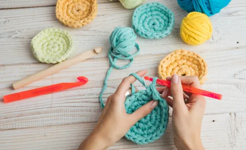Hands crocheting a round item, balls of colorful yarn around them.