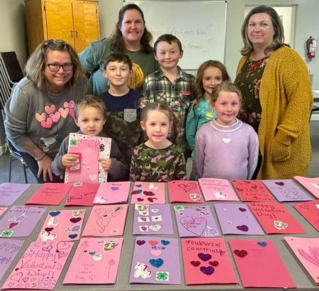 A group of six children and three adults stand behind a table covered in colorful, handmade Valentine's Day cards. The children are smiling and some wear 4-H themed apparel. The decorated cards are in shades of pink, red, and purple.