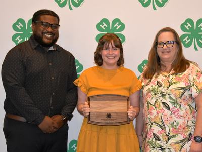 Three people pose for a photo at a 4-H awards event, with the center person holding an award plaque. The group stands in front of a backdrop featuring green 4-H logos.