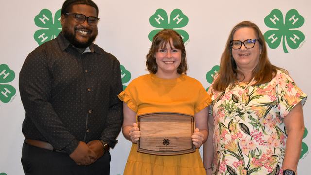 Three people pose for a photo at a 4-H awards event, with the center person holding an award plaque. The group stands in front of a backdrop featuring green 4-H logos.