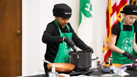 4-H member wearing a green apron and black chef hat stirring a pot.