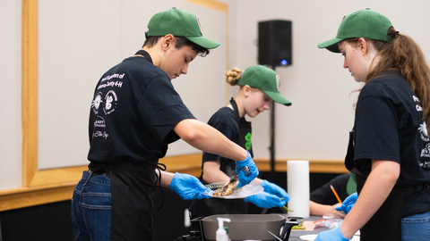 Three 4-H members in green caps, black shirts and blue gloves work at a cooking station; one uses tongs to plate food while others assist.