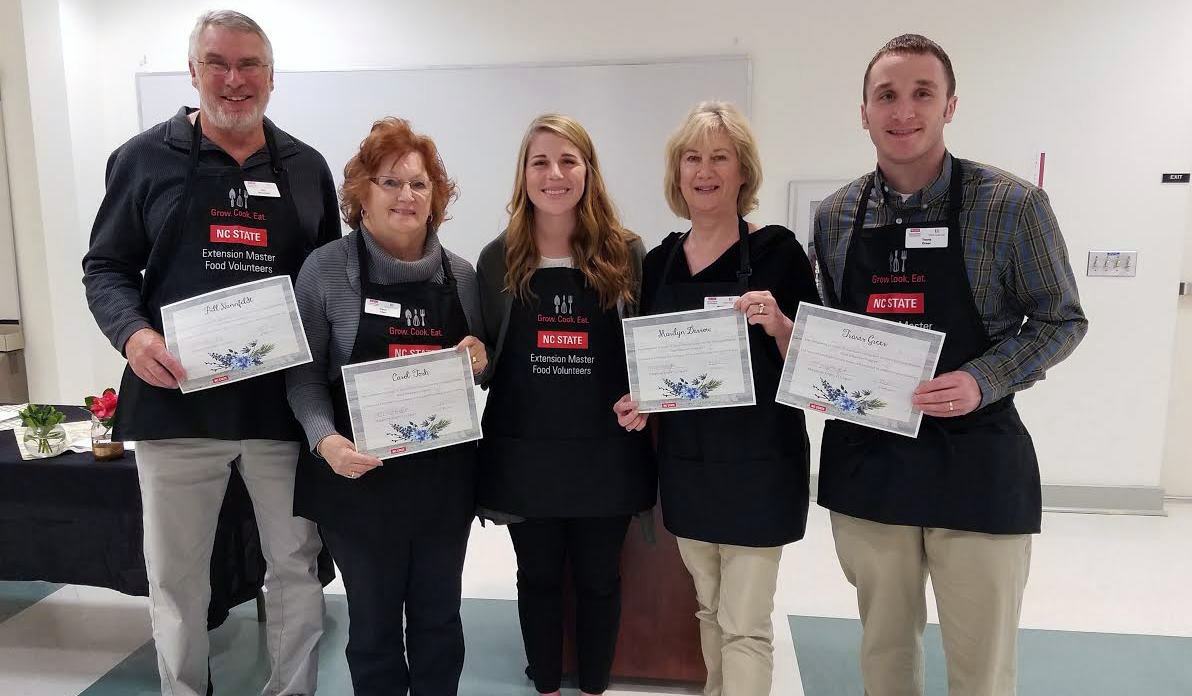 Volunteers wearing black aprons show their certificates after being trained to serve in the NC State Extension Master Food Volunteers program.