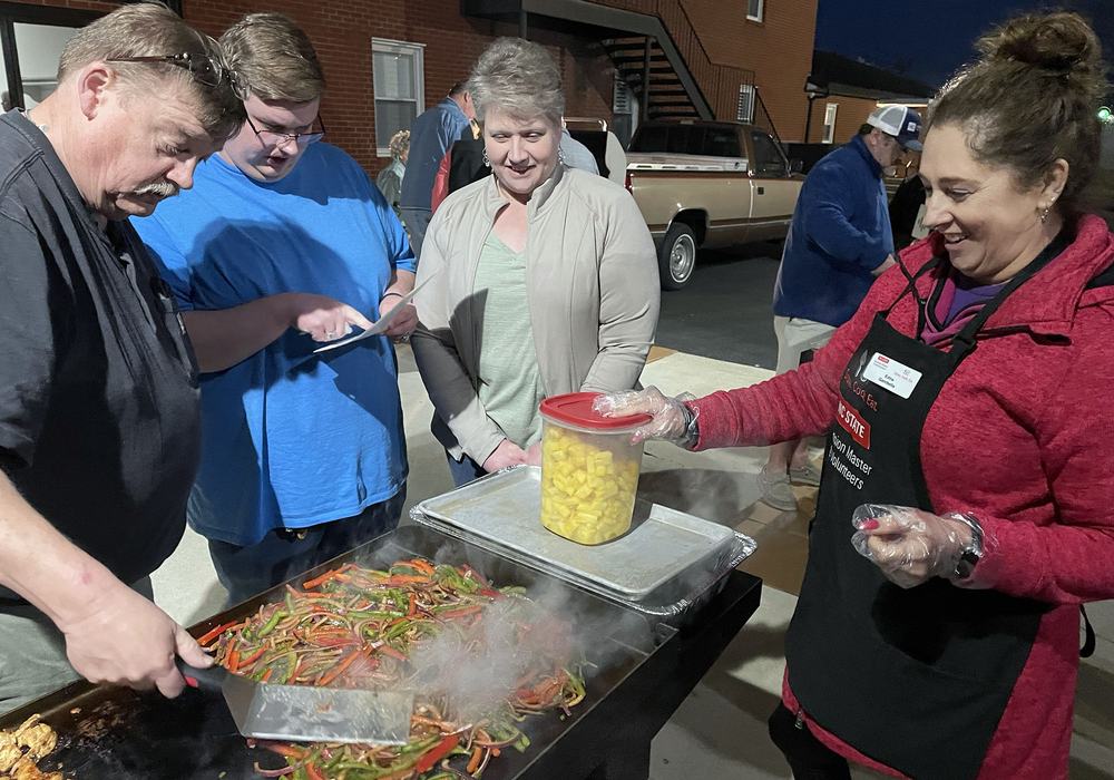 An NC State Extension Master Food Volunteer program volunteer wearing a black apron with the program logo conducts a cooking demonstration as three people look on