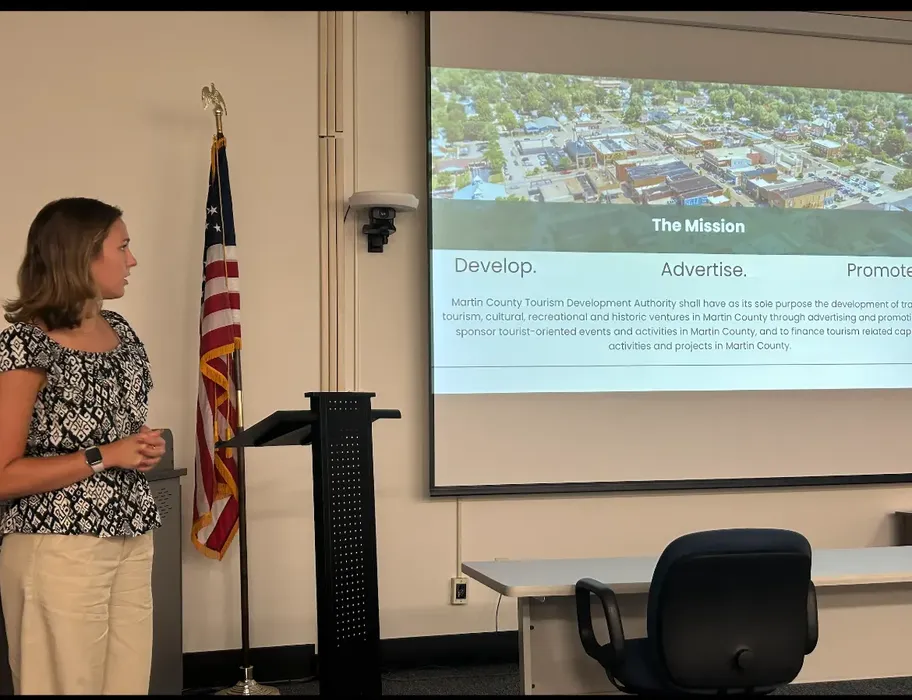 A young woman (Emily) in a flower blouse and slacks is standing in a conference room to the left side of a slide show presentation showing an overhead view of a town on top and text below.