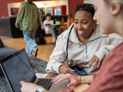 Two female college students look at a laptop screen in a campus building.