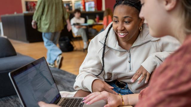 Two female college students look at a laptop screen in a campus building.
