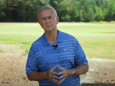 A person in a blue striped polo stands in an open, grassy field with trees in the background, preparing to speak.
