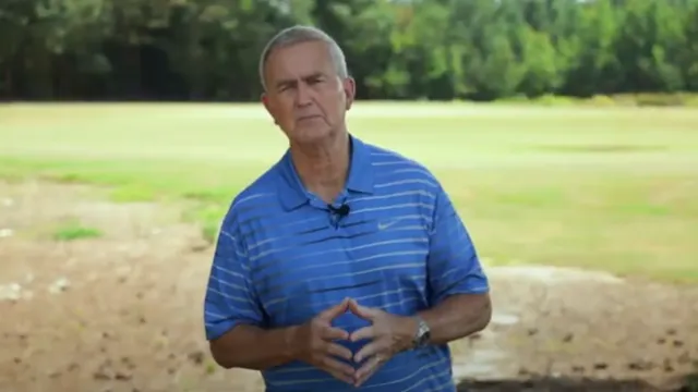 A person in a blue striped polo stands in an open, grassy field with trees in the background, preparing to speak.