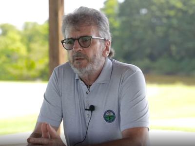 Man wearing glasses and a gray polo with a lapel mic speaks outdoors, appearing thoughtful against a sunny, leafy background.