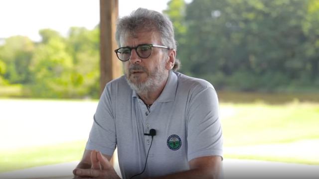 Man wearing glasses and a gray polo with a lapel mic speaks outdoors, appearing thoughtful against a sunny, leafy background.
