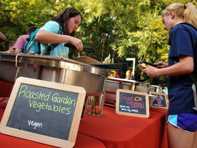 Woman browsing the local cooked foods at a farmers market. Featuring Roasted Garden Vegetables labeled by a sign.