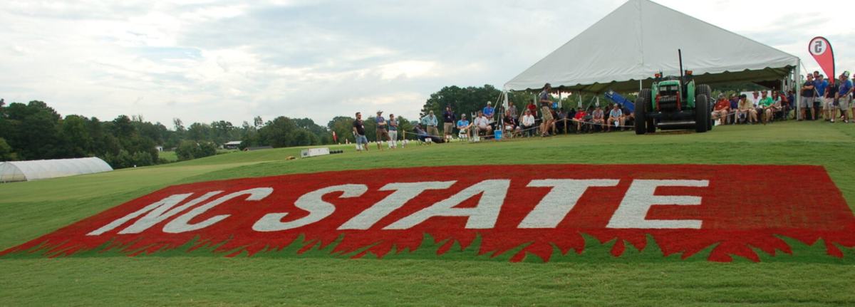 Red NC State Logo is spray painted on the grass of a hill. At the top of the hill is a pavilion and a tractor with people gathered around them. The sky is blue and lightly clouded.