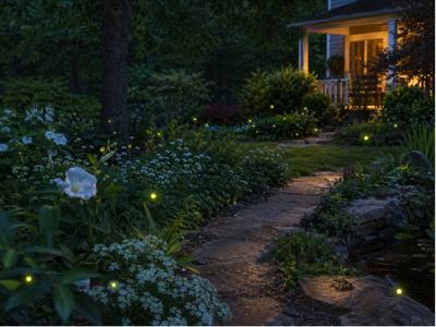 A garden at nighttime with light colored flowers, fireflies illuminating their surroundings, and a walkway leading to the house's front porch with the light on.