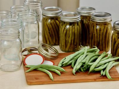 seven empty canning jars and lids beside five jars of canned green beans with fresh picked green beans laying on a cutting board.