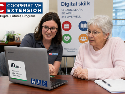 A staff member helps an older woman at a laptop labeled "ID.me." NC Cooperative Extension logo and a "Digital skills" poster are visible, suggesting a digital skills training session.