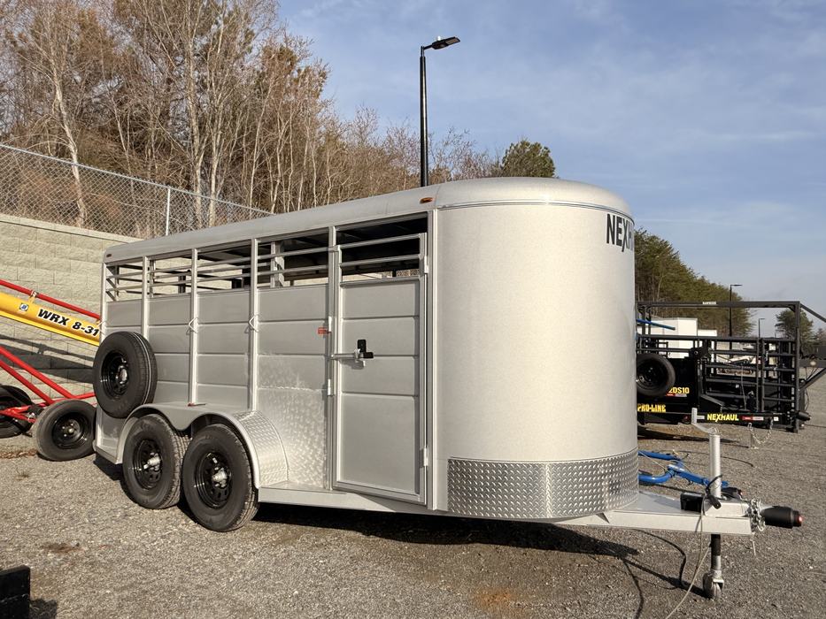 A silver, tandem-axle NexHaul livestock trailer with a rounded nose and slatted sides for ventilation. It features black wheels, a side access door, and a spare tire mounted on the rear, parked on a gravel lot under a clear blue sky.