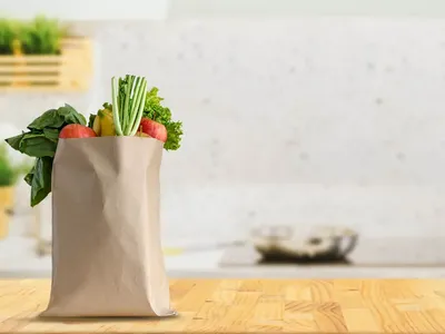 A paper grocery bag sits atop at a kitchen county overflowing with fresh produces and healthy foods while a stovetop and cooking utensils are seen in the background.