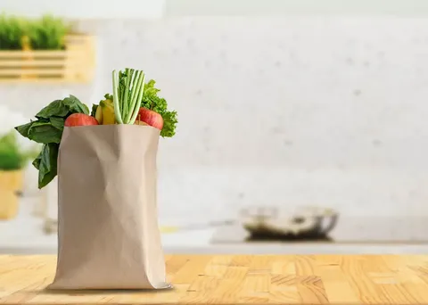 A paper grocery bag sits atop at a kitchen county overflowing with fresh produces and healthy foods while a stovetop and cooking utensils are seen in the background.