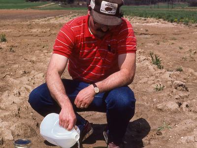 Person pouring liquid into pan in agricultural field