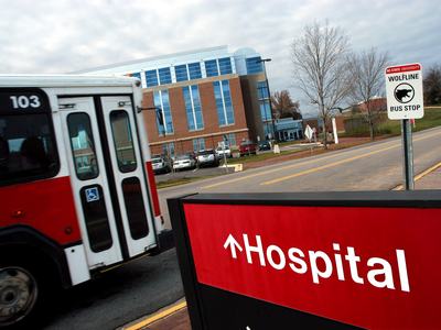 Red sign with "Hospital" painted in white letters. A red and white bus and hospital building in the background.