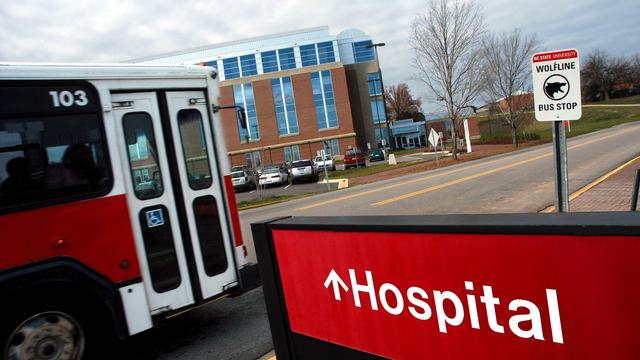 Red sign with "Hospital" painted in white letters. A red and white bus and hospital building in the background.