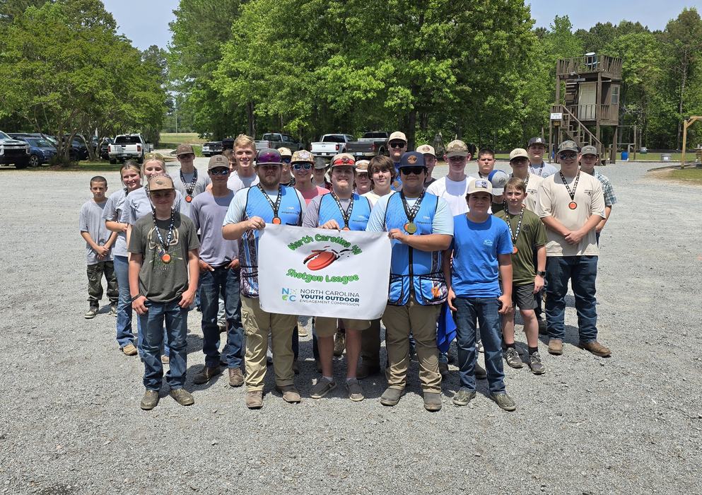 A group of youths and teens wearing casual outdoor clothing and caps stand together on a gravel area, many with medals around their necks.  Several in front hold a banner reading “North Carolina 4-H Shotgun League” with a clay target graphic.