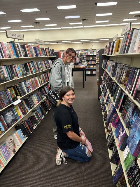 In between the bookshelves in a bookstore, two girls are shopping for books. One girl kneels with a book on her lap. Another girl stands next to her, on the far side. Both girls are looking toward the camera with quiet smiles.