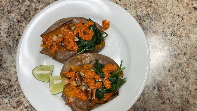 Cooked sweet potatoes with chickpeas, spinach, and red pepper sauce with two pieces of lime all on a white dinner plate on a kitchen counter.