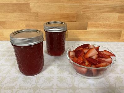 Clear glass bowl of strawberries with two jars of strawberry jam next to it on a kitchen table with a wooden background.