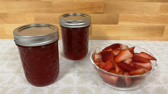 Clear glass bowl of strawberries with two jars of strawberry jam next to it on a kitchen table with a wooden background.