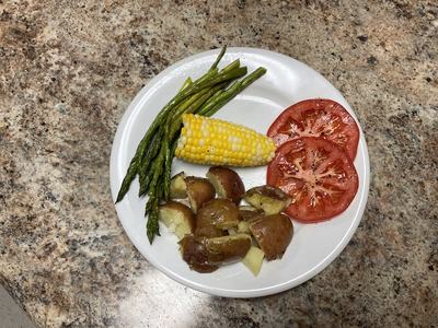 White dinner plate with roasted asparagus, corn on the cob, boiled red skinned potatoes, and sliced tomatoes.